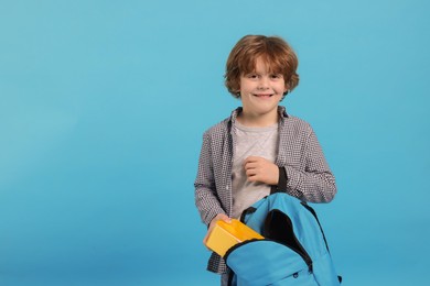 Little boy packing lunch box into backpack on light blue background. Space for text