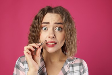 Photo of Emotional woman plucking her mustache with tweezers on pink background