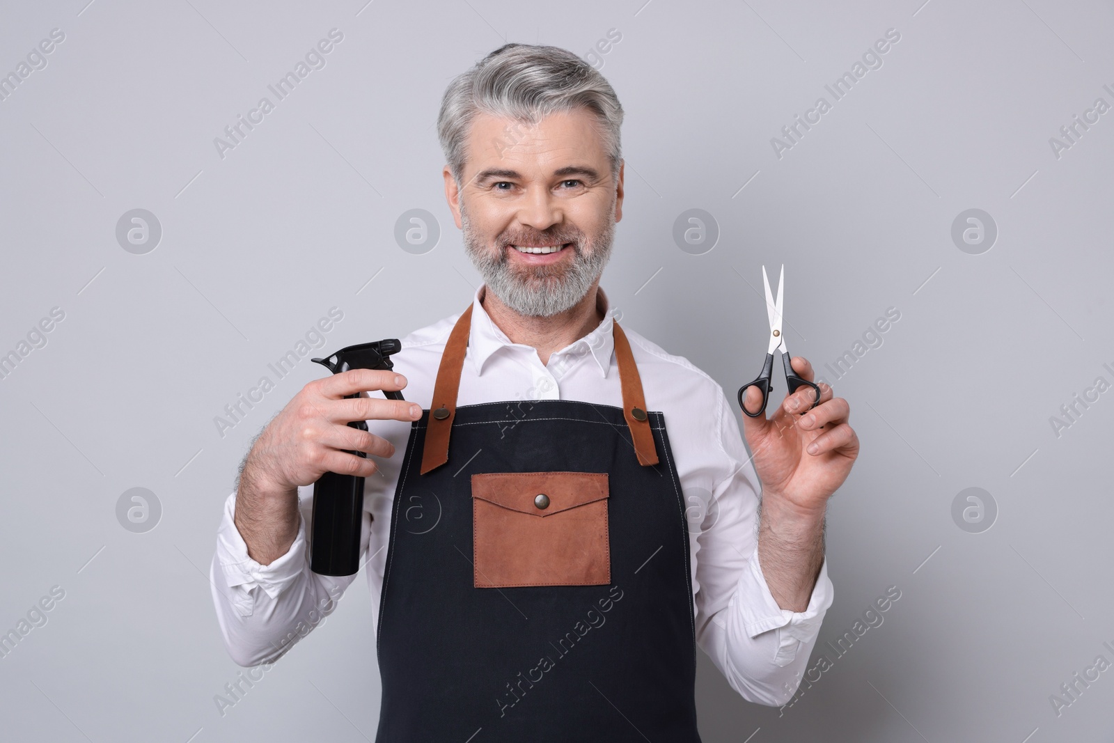 Photo of Smiling hairdresser with scissors and spray bottle on gray background