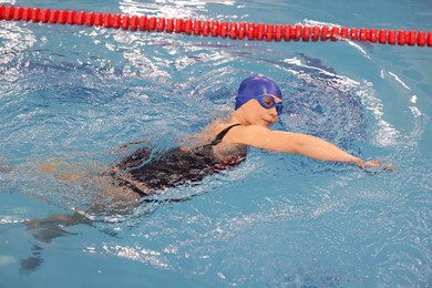 Sporty woman wearing cap and goggles swimming in pool indoors