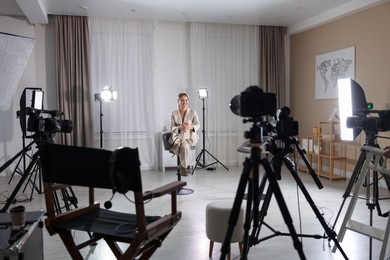 Photo of Young woman sitting on chair near cameras and other professional equipment in modern film studio