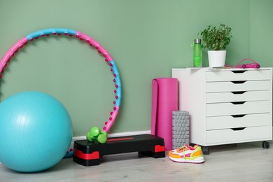 Photo of Different fitness equipment, sneakers and chest of drawers near green wall indoors