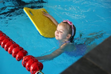 Photo of Little girl with goggles and kickboard swimming in pool indoors