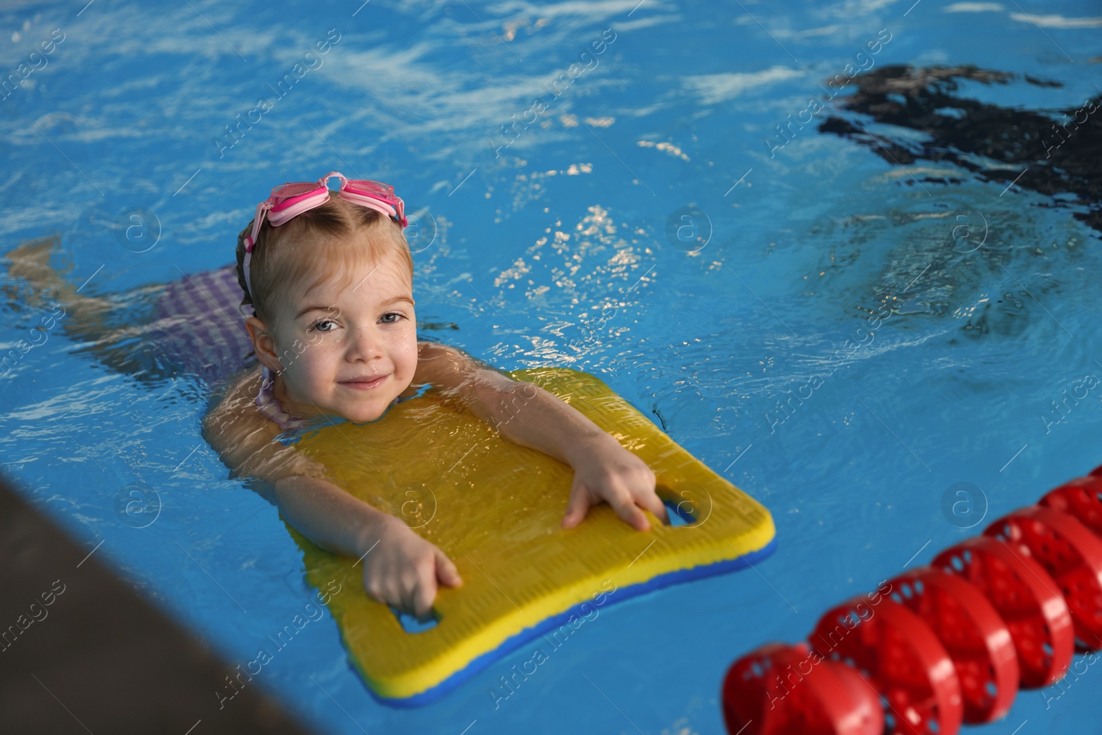 Photo of Little girl with goggles and kickboard swimming in pool indoors