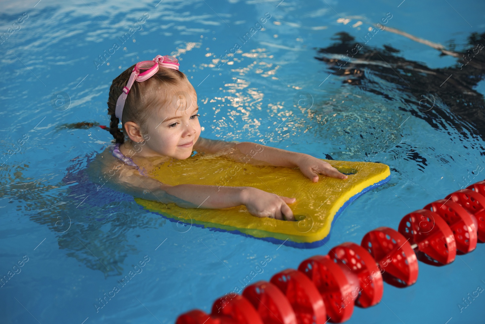 Photo of Little girl with goggles and kickboard swimming in pool indoors