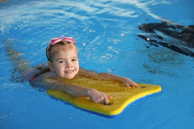 Photo of Little girl with goggles and kickboard swimming in pool indoors