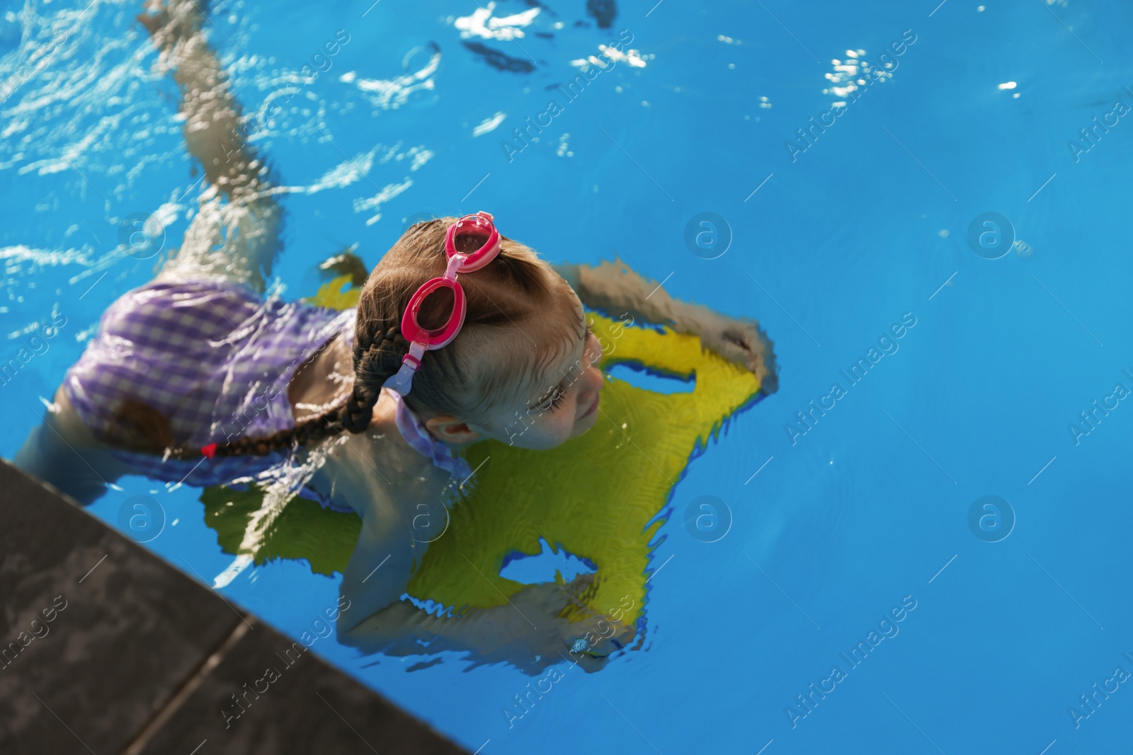 Photo of Little girl with goggles and kickboard swimming in pool indoors, above view