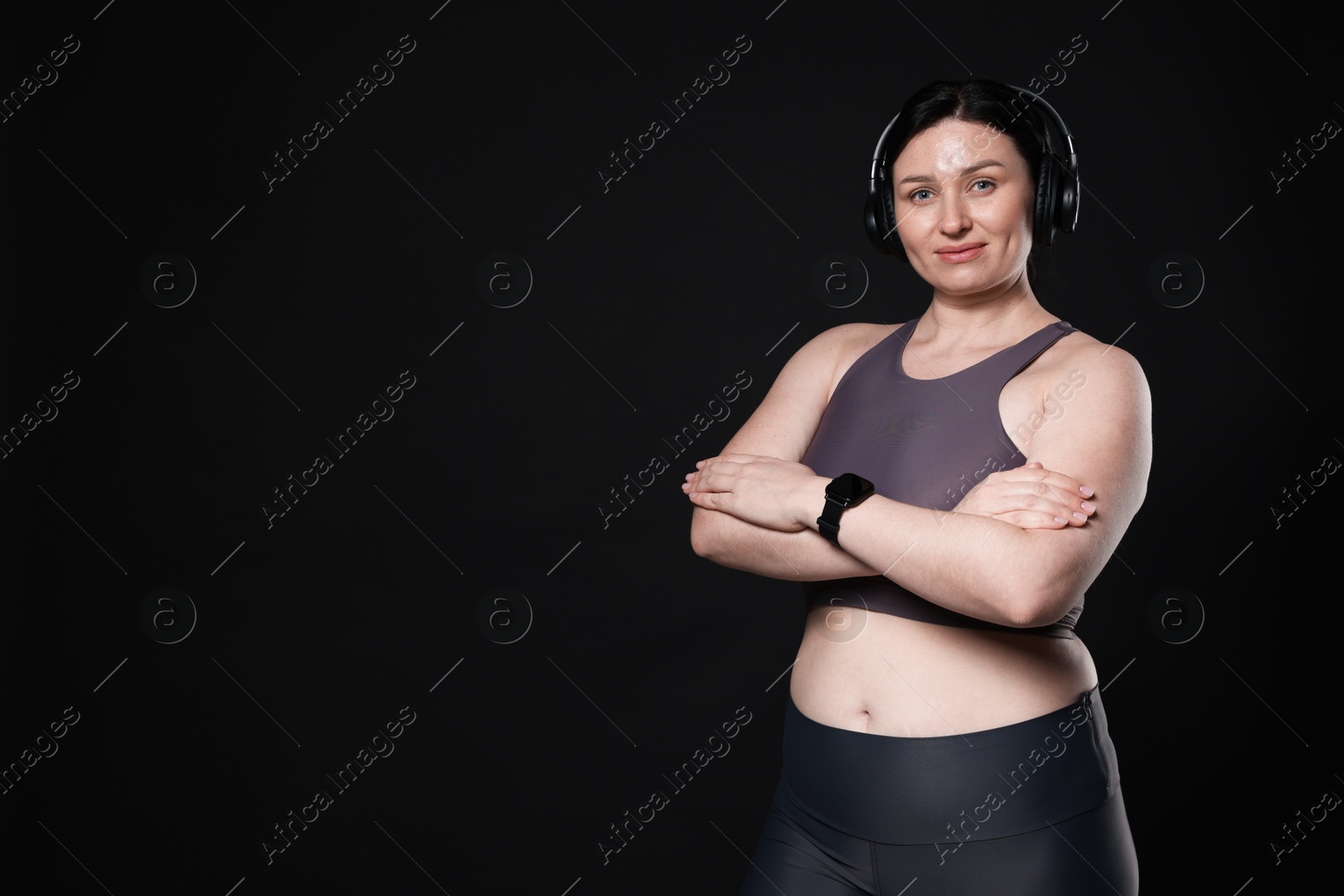 Photo of Plus size woman in gym clothes and headphones on black background, space for text