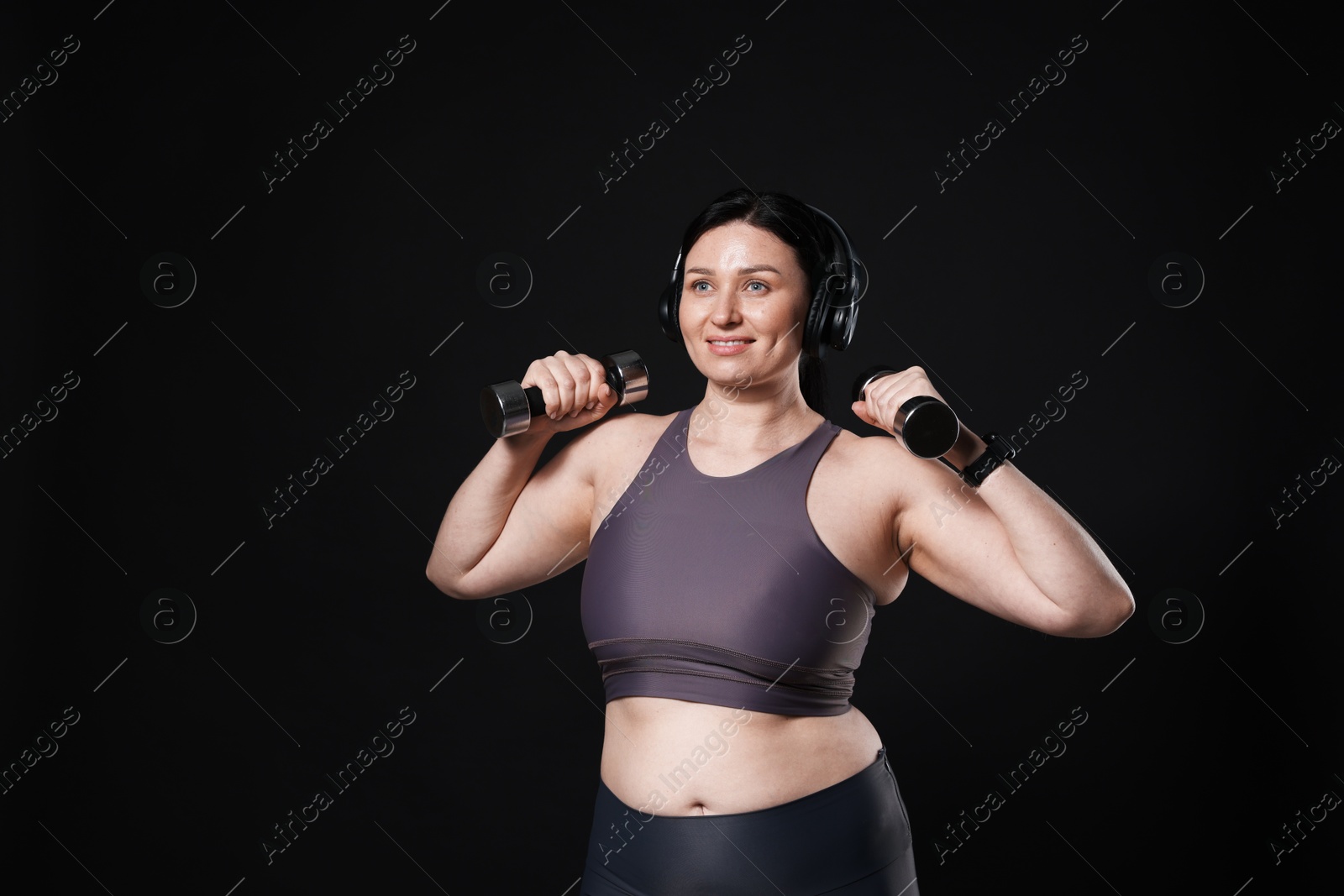 Photo of Plus size woman in gym clothes with headphones and dumbbells on black background