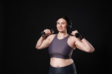 Plus size woman in gym clothes with headphones and dumbbells on black background