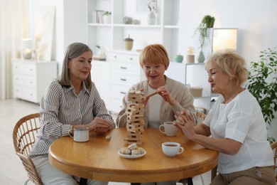 Photo of Friendship. Senior women building tower with wooden blocks at wooden table indoors