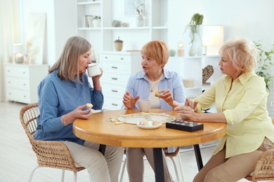 Friendship. Senior women enjoying hot drinks and cookies at table indoors