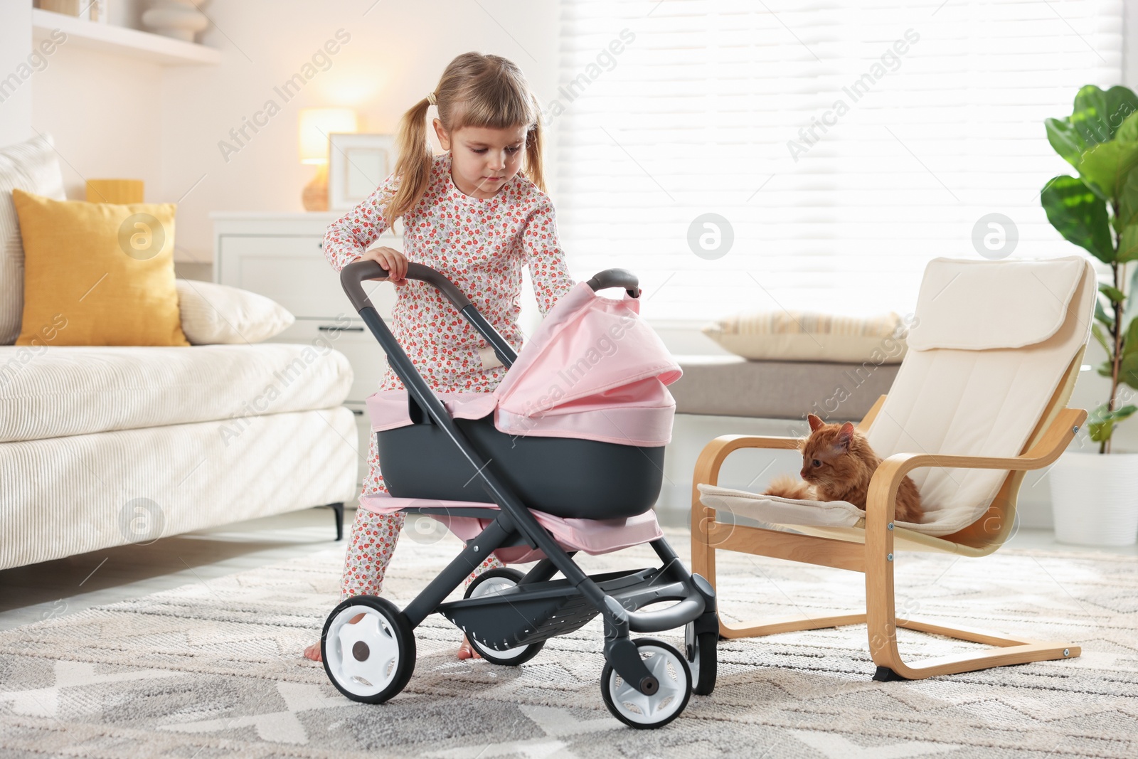 Photo of Cute little girl with doll stroller and ginger cat sitting in armchair at home