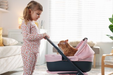 Photo of Cute little girl with her ginger cat in doll stroller at home