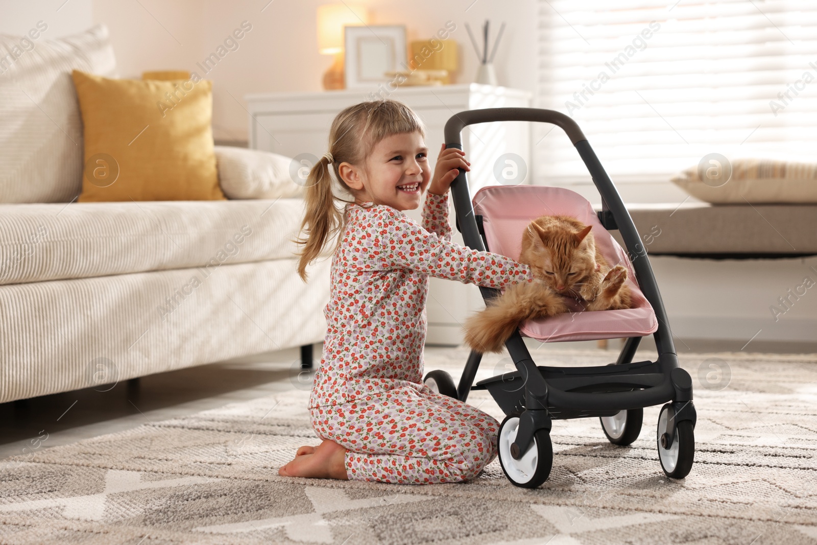 Photo of Cute little girl with her ginger cat in doll stroller at home