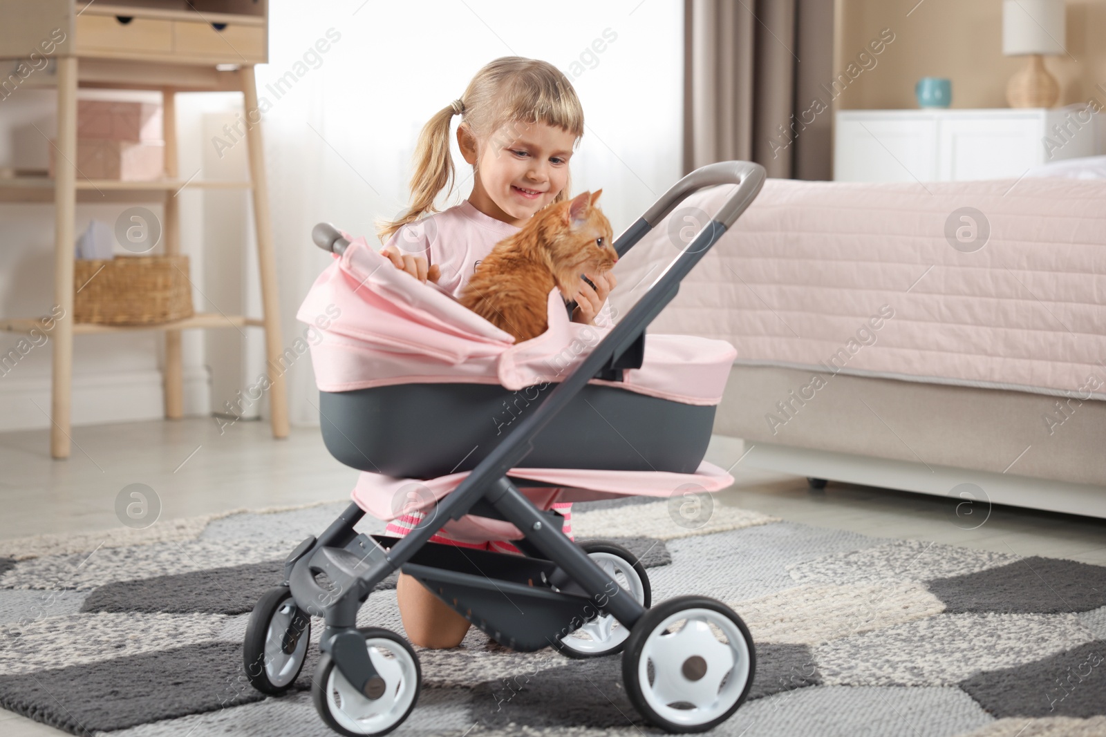 Photo of Cute little girl with her ginger cat in doll stroller at home