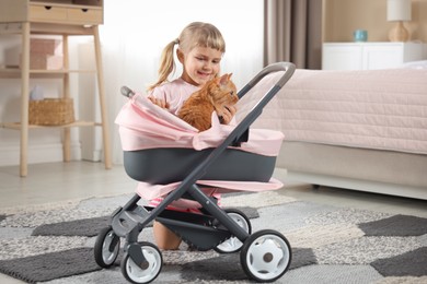 Photo of Cute little girl with her ginger cat in doll stroller at home