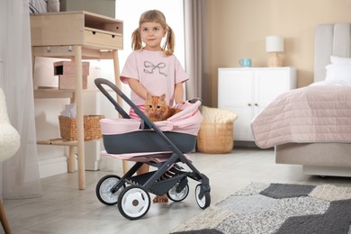 Photo of Cute little girl with her ginger cat in doll stroller at home