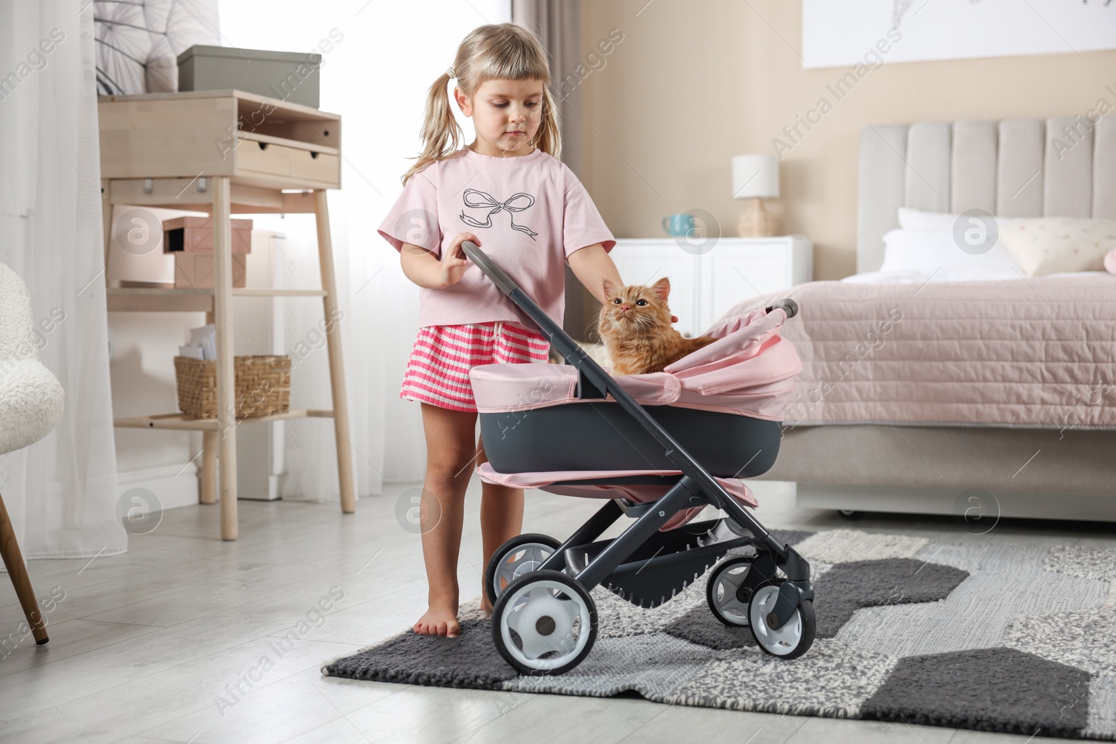 Photo of Cute little girl with her ginger cat in doll stroller at home