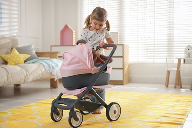 Photo of Cute little girl putting teddy bear into doll stroller at home