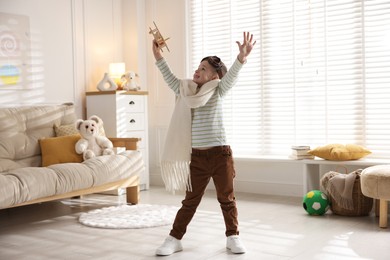 Photo of Happy little boy in scarf and aviator goggles playing with toy plane at home