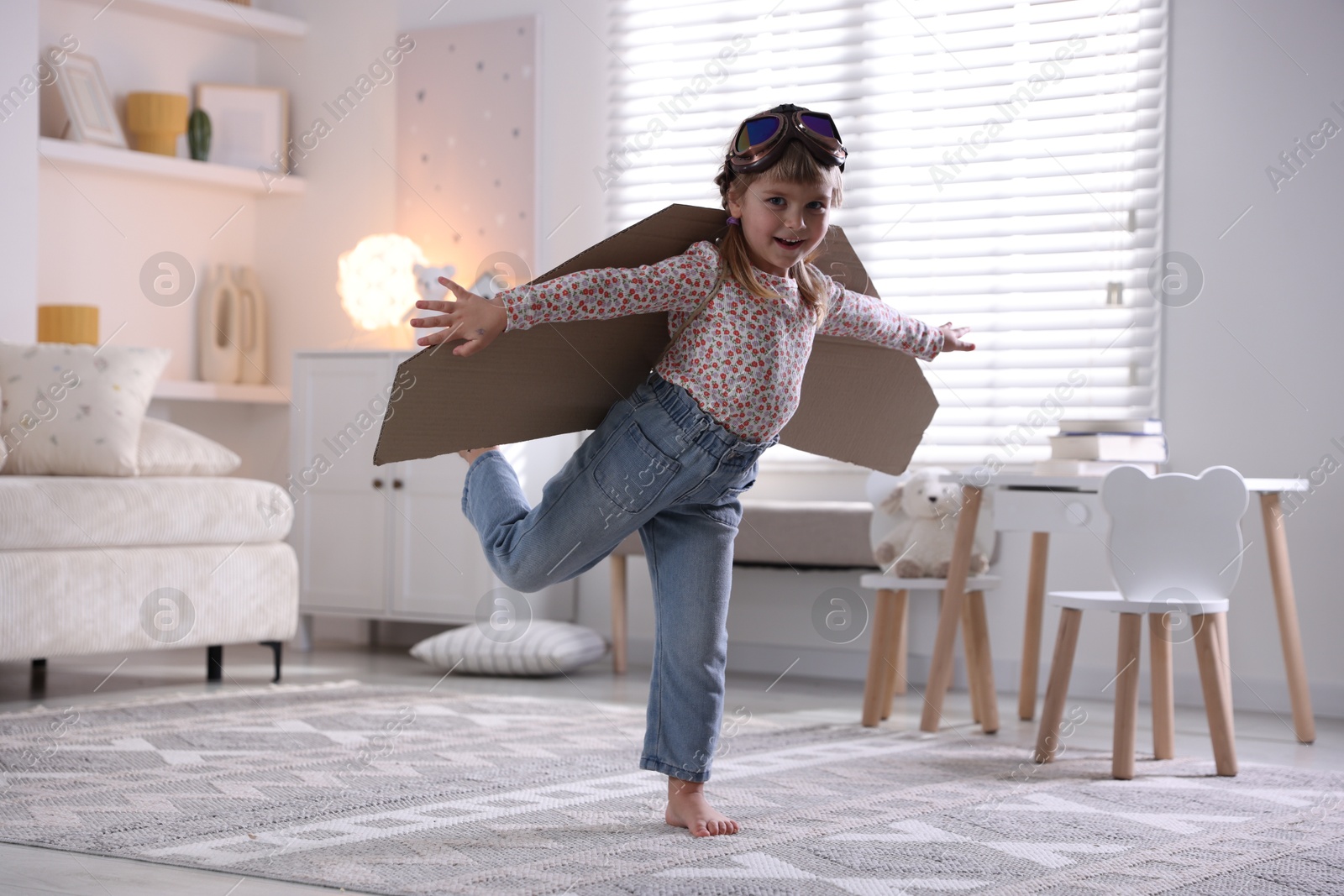 Photo of Cute little girl with cardboard plane wings and goggles playing pilot at home