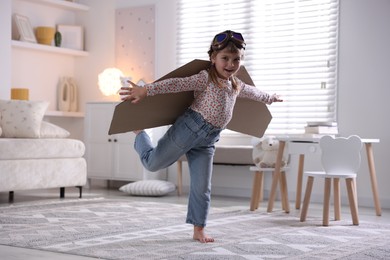 Cute little girl with cardboard plane wings and goggles playing pilot at home