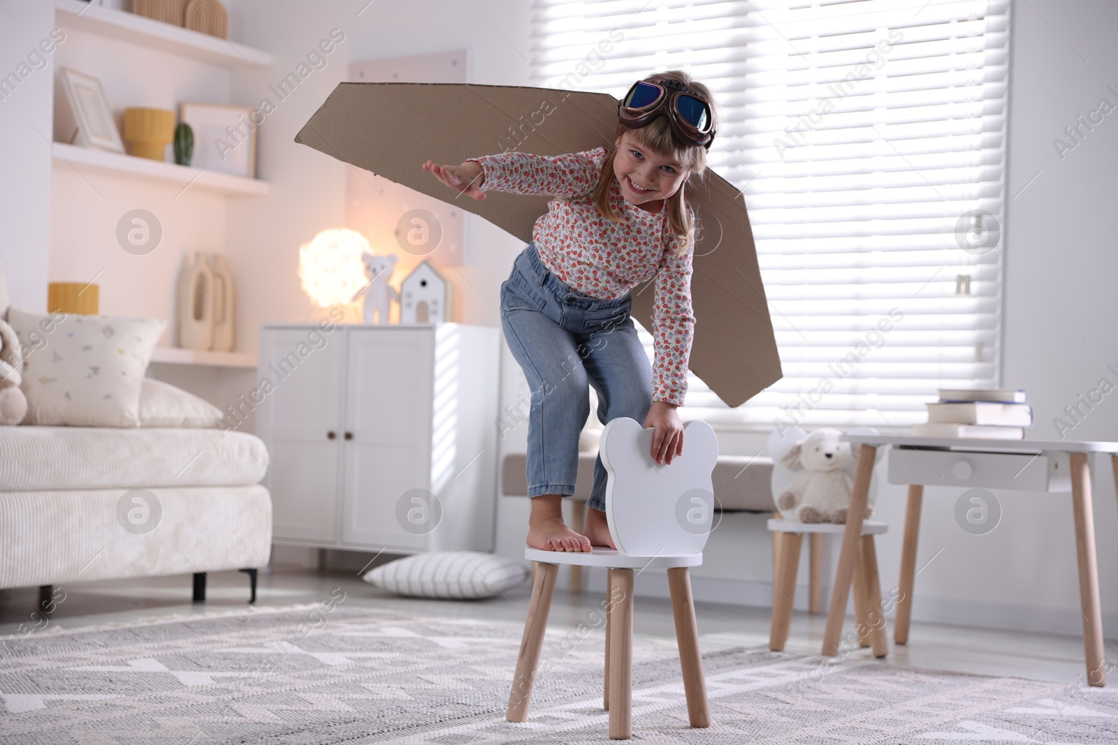 Photo of Cute little girl with cardboard plane wings and goggles playing pilot at home