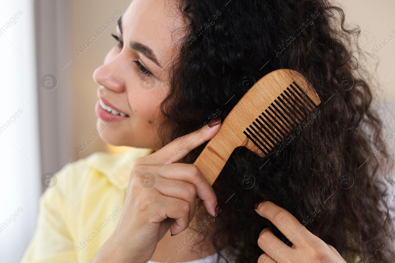 Photo of Smiling young woman brushing her curly hair with comb at home, closeup