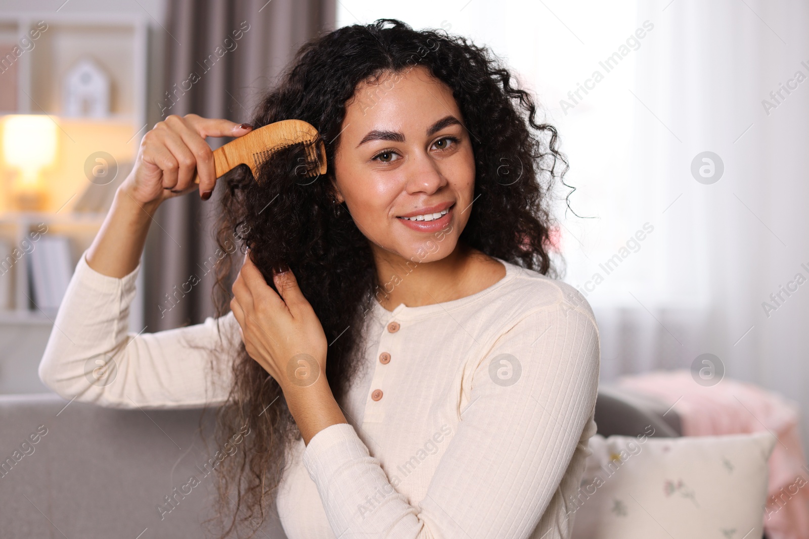 Photo of Smiling young woman brushing her curly hair with comb at home