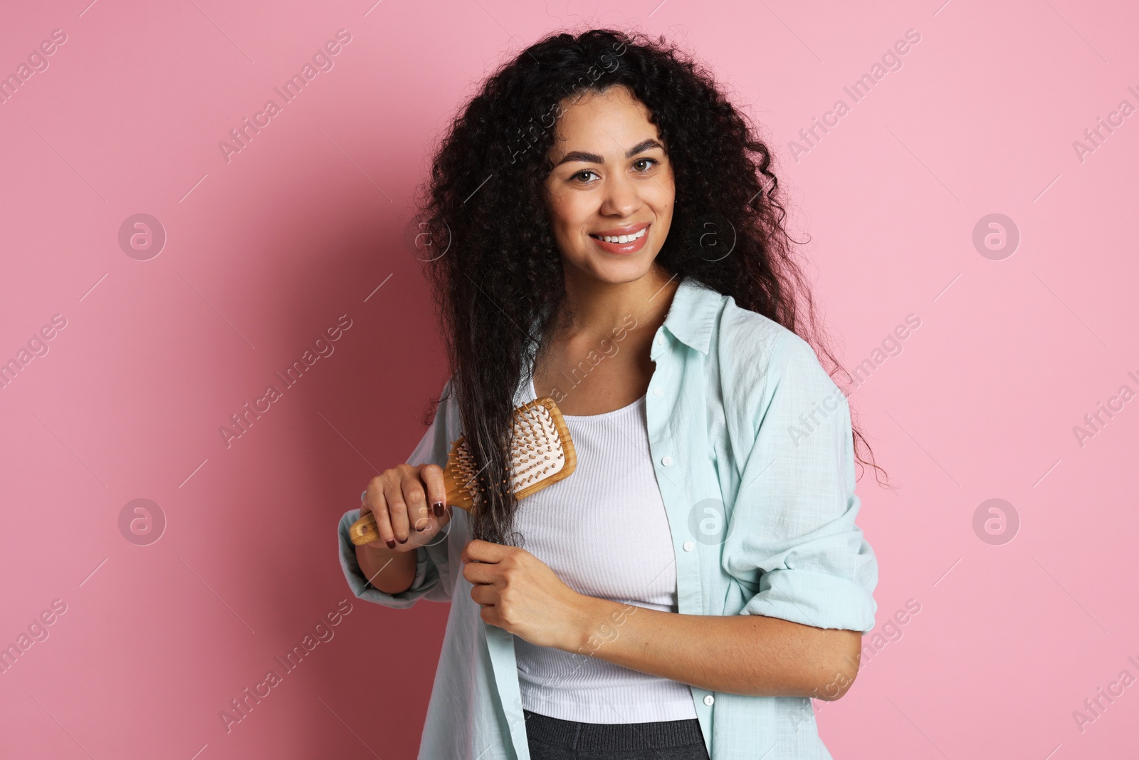 Photo of Smiling young woman brushing her curly hair on pink background