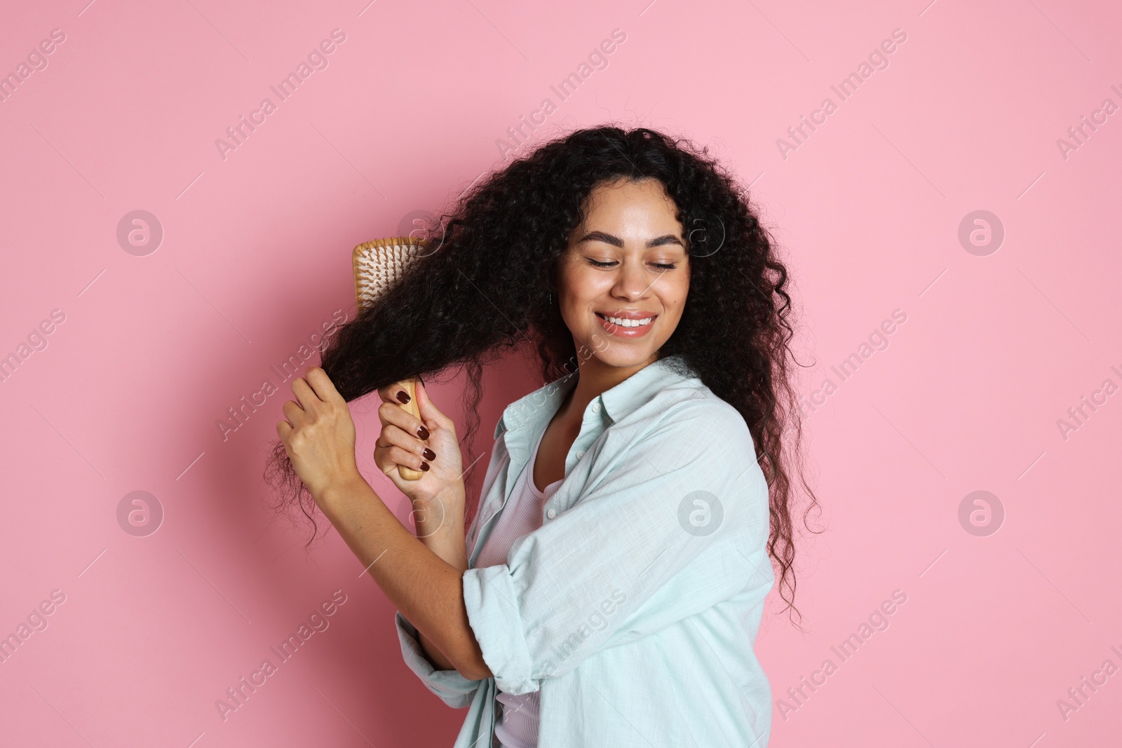 Photo of Smiling young woman brushing her curly hair on pink background