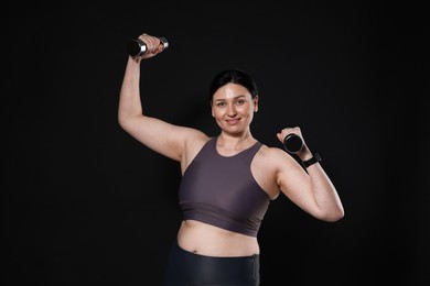 Plus size woman in gym clothes doing exercise with dumbbells on black background