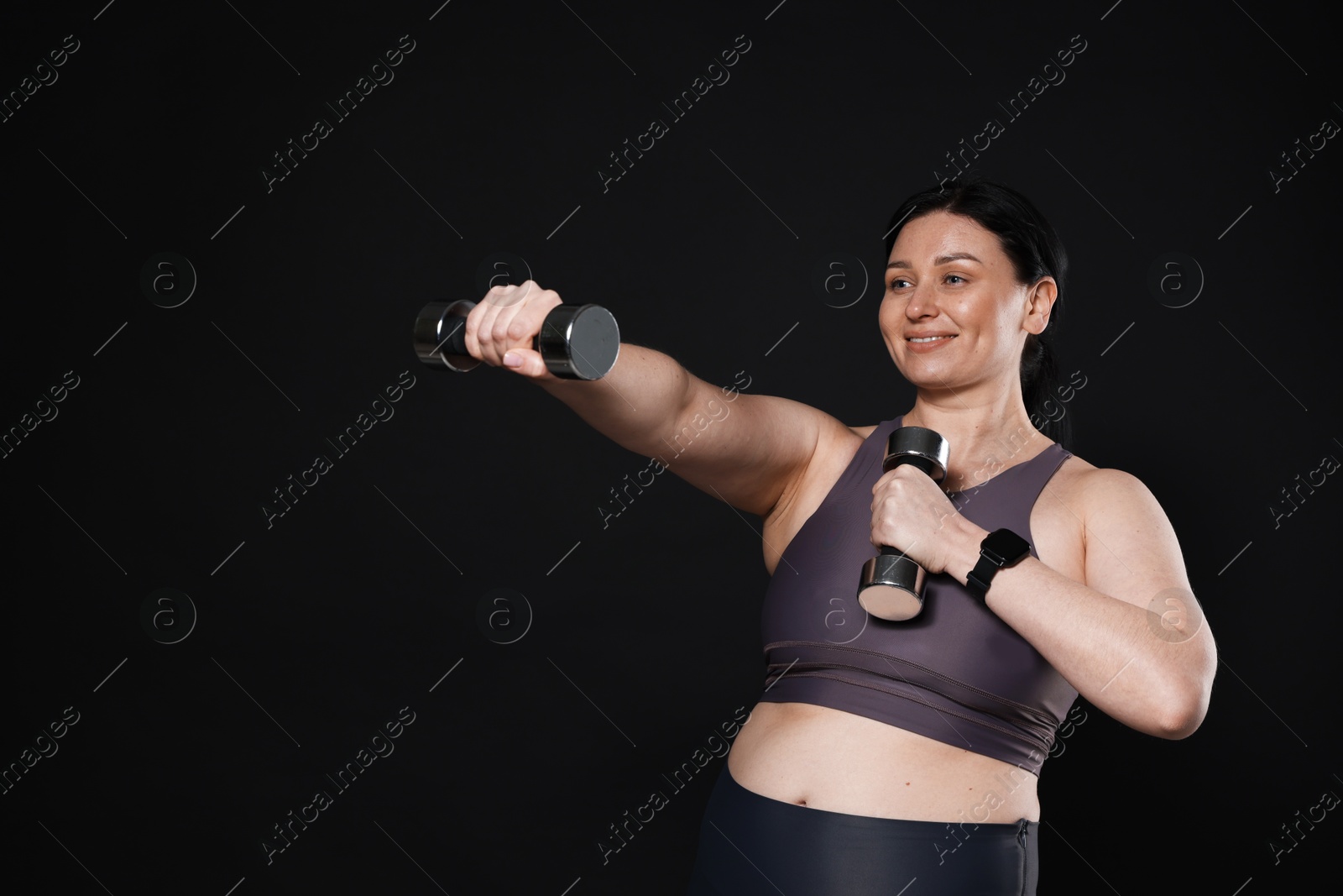 Photo of Plus size woman in gym clothes doing exercise with dumbbells on black background, space for text