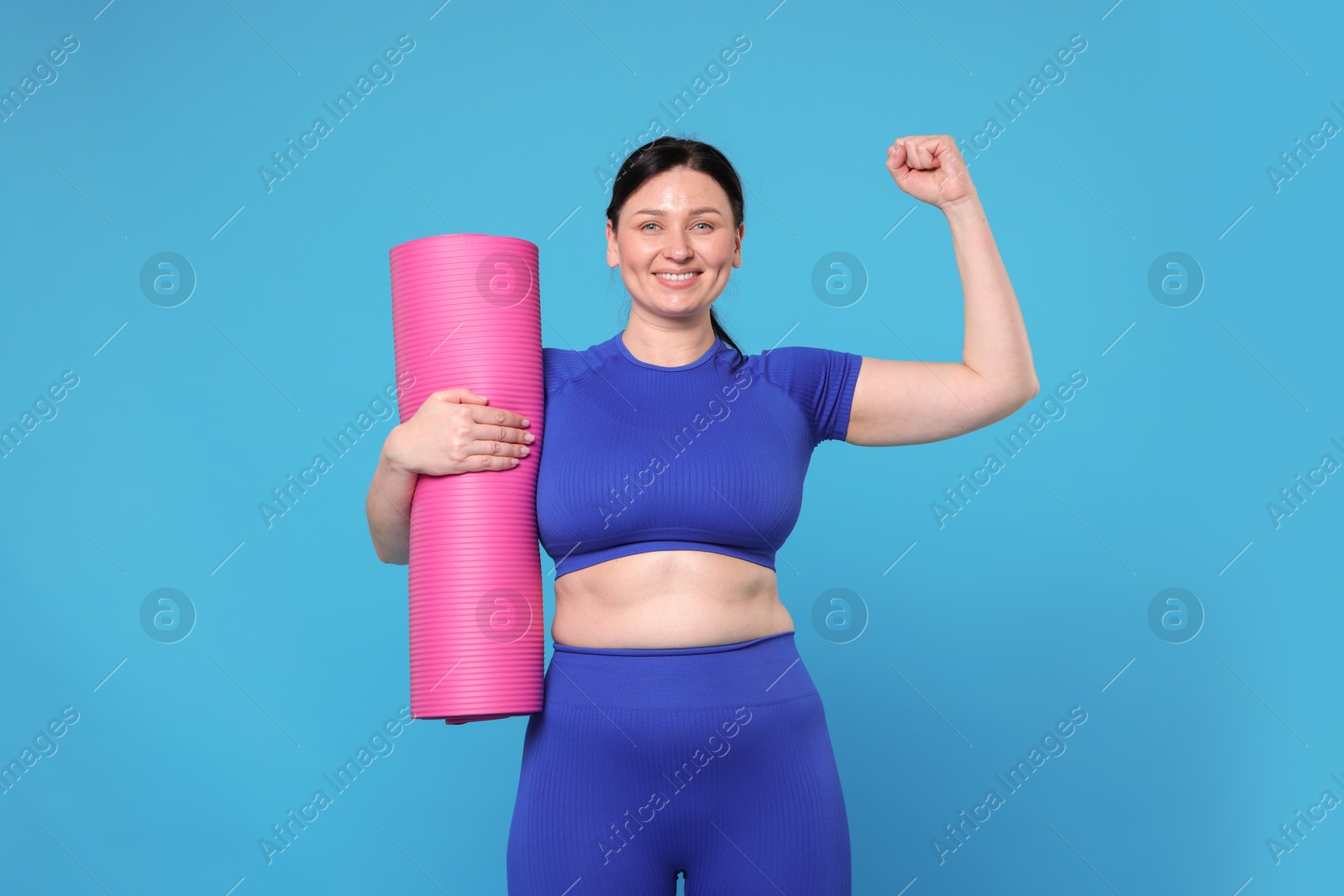 Photo of Plus size woman in gym clothes with fitness mat showing her bicep on light blue background