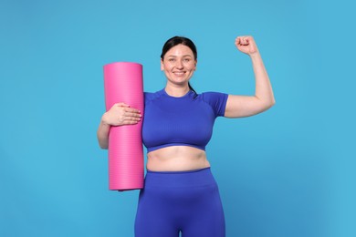 Plus size woman in gym clothes with fitness mat showing her bicep on light blue background