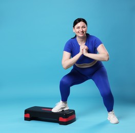 Plus size woman in gym clothes doing exercise with step platform on light blue background