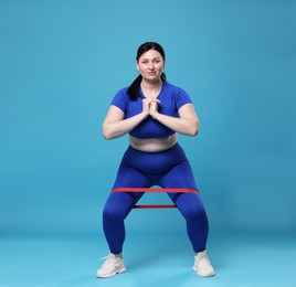 Plus size woman in gym clothes doing exercise with elastic band on light blue background