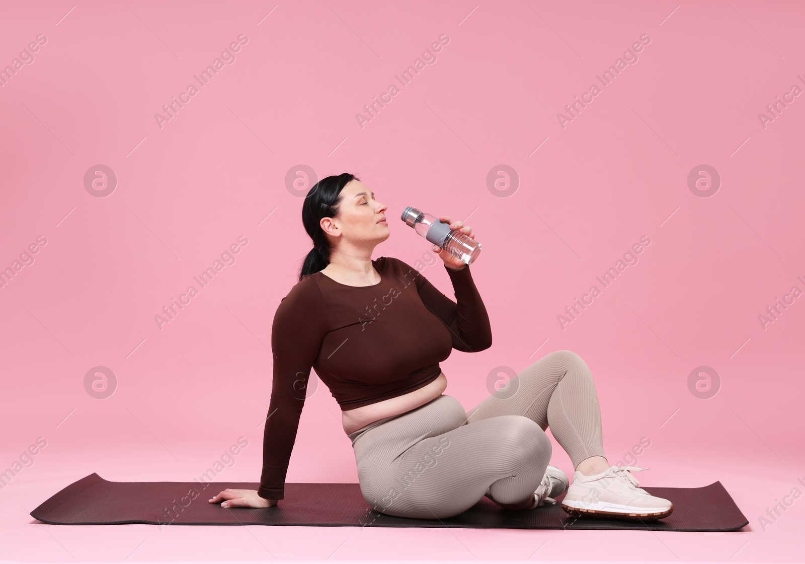 Photo of Plus size woman in sportswear drinking water on pink background