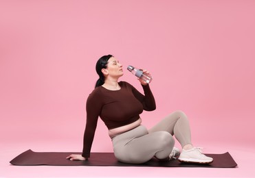 Plus size woman in sportswear drinking water on pink background