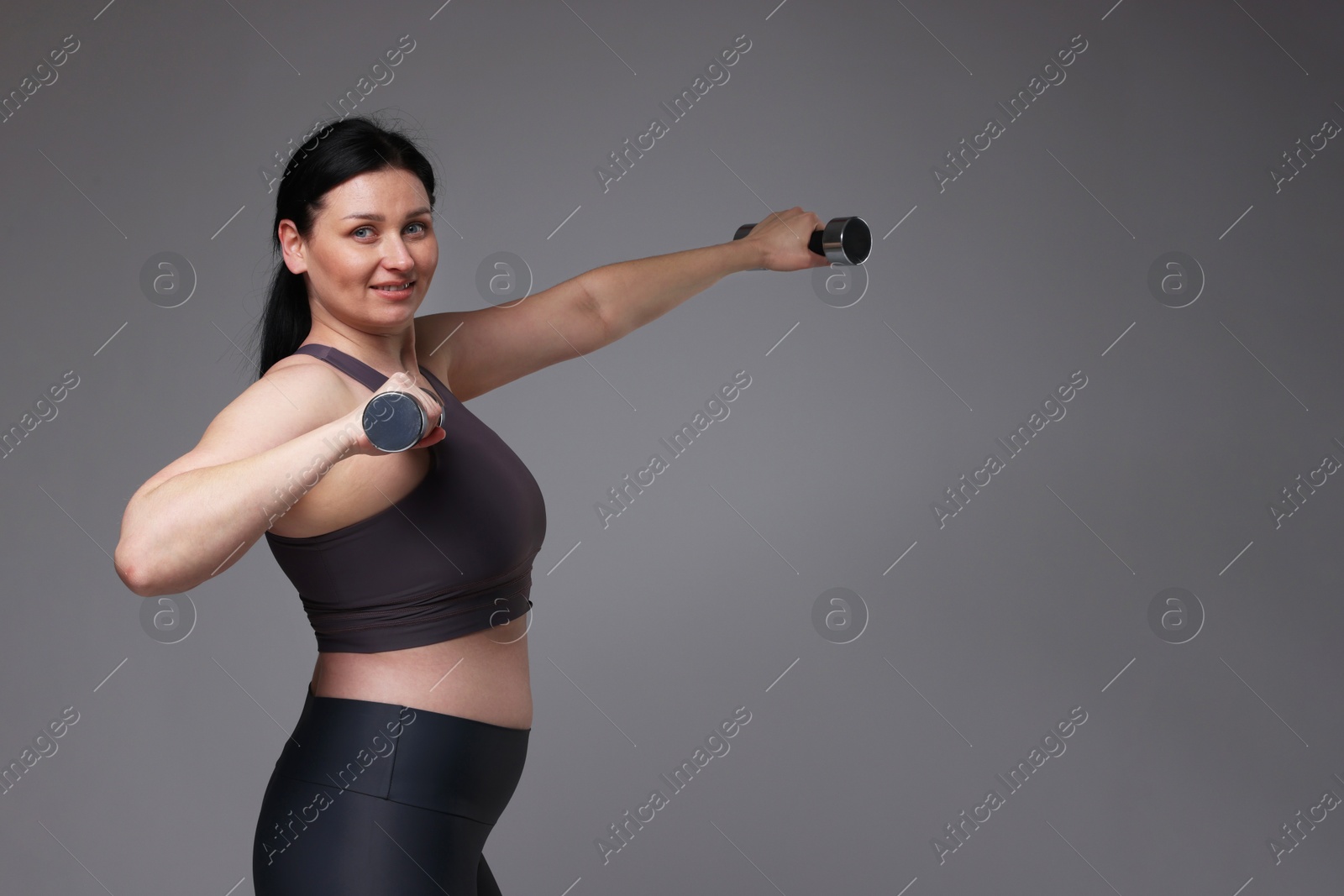 Photo of Plus size woman in gym clothes doing exercise with dumbbells on grey background, space for text