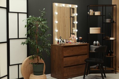 Photo of Makeup room. Wooden dresser with mirror, different beauty products, shoes, houseplant and chair indoors