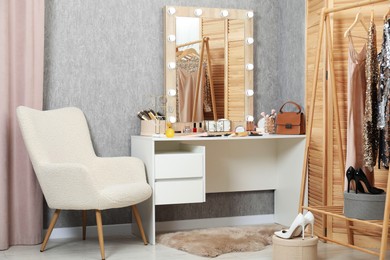 Photo of Makeup room. Mirror, dressing table, different beauty products, clothing rack and armchair indoors