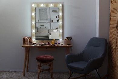 Photo of Makeup room. Mirror, wooden dressing table, different beauty products, stool and armchair indoors