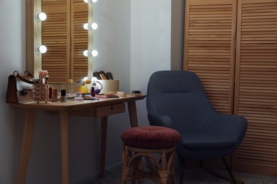 Photo of Makeup room. Mirror, wooden dressing table, different beauty products, stool and armchair indoors