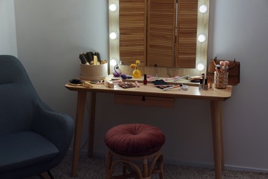 Photo of Makeup room. Mirror, wooden dressing table, different beauty products, stool and armchair indoors