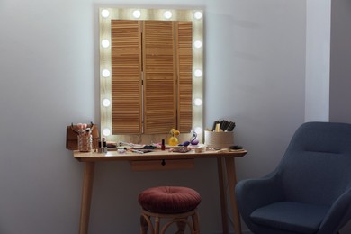 Photo of Makeup room. Mirror, wooden dressing table, different beauty products, stool and armchair indoors