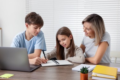 Photo of Smiling mother helping her children with homework at table indoors