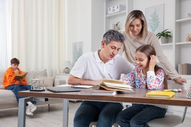 Photo of Parents helping their daughter with homework while son reading book on sofa indoors