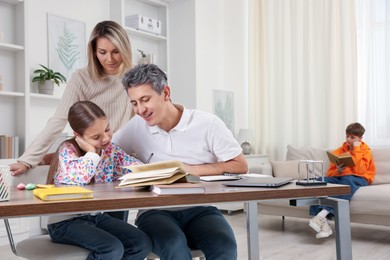 Photo of Parents helping their daughter with homework while son reading book on sofa indoors